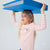 A young girl with blonde hair wearing a pink and white striped long-sleeve shirt and pink hair clips, holding a large blue binder over her head against a light gray background.