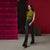 A woman models a dark olive green sleeveless top, black flared trousers, and black loafers, carrying a dark brown leather crossbody bag. She stands in a studio setting with a deep magenta velvet panel on the left and a solid dark red wall on the right.