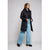 A woman modeling a long black puffer jacket, styled with a black top, wide-leg light blue jeans, and black boots, against a plain light gray studio background.