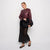 A woman models a burgundy faux-leather moto jacket with black mesh sleeves, paired with a black skirt and holding a black jacket, against a plain white studio background.