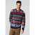 A man wearing a navy blue Christmas sweater with festive patterns of reindeer, gingerbread men, candy canes, and 'MERRY XMAS' text, paired with brown pants, against a light gray background.