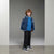 A young boy with curly brown hair stands in a studio wearing a navy blue varsity-style jacket with white stripes, a bright blue t-shirt, dark jeans, and navy sneakers.