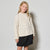 A young girl with blonde hair stands against a light gray studio background wearing a cream cable-knit mock-neck sweater and a black skirt.