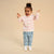 A toddler girl with her hair in a high bun stands smiling with arms raised, wearing a pink and white striped long-sleeve top, light blue jeans, and white sneakers against a solid beige studio background.