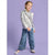 A young girl modeling a shiny silver faux-leather jacket, styled with a white patterned shirt, wide-leg blue jeans, and silver flats, set against a solid lavender studio background.