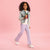 A young girl with curly black hair is modeling a light-wash denim jacket, a white t-shirt with a red cherry graphic, lavender flare-leg pants, and white sneakers, set against a solid pink studio background.