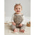 A smiling baby girl sitting on a cream knit blanket, wearing a beige and black leopard print overall romper over a white long-sleeve shirt. She has two small hair bows and is posed in a bright, neutral studio setting with a small woven basket to her side.