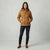 A woman modeling a tan hooded puffer jacket, paired with dark blue skinny jeans and brown ankle boots, standing against a light gray studio background.