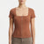 A woman wearing a fitted short-sleeve top in a warm terracotta or rust brown color, featuring a square neckline and a front zipper closure. The image is cropped from the lower face to the hips against a clean white studio background.
