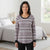 A woman wearing a long-sleeve crewneck top with a black, white, and pink Fair Isle pattern, standing in a brightly lit bedroom.