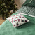 A festive throw pillow with a red Christmas truck and tree pattern resting on a green snowflake-patterned blanket, with a blurred Christmas tree in the background.