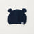 A navy blue knit baby beanie hat with two pom-pom ears on top and a small white square patch with a black paw print near the brim, shown on a plain off-white background.
