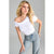 A blonde woman posing in a white off-the-shoulder short-sleeve top and light-wash blue jeans against a solid gray studio background.