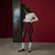 A model stands in a studio setting wearing a white textured long-sleeve top, a pleated burgundy mini skirt, black tights, white socks, and brown sneakers. The background features a deep red wall, a dark vertical panel, and a concrete floor.