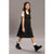 A young girl wearing a black sleeveless dress with a small white floral pattern over a white t-shirt, paired with white socks and black ankle boots, walking against a solid gray studio background.