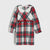 A long-sleeve children's dress with a red, green, and white tartan plaid pattern, featuring a solid white Peter Pan collar and a matching red waist tie, shown against a light gray background.