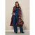 A woman models a long burgundy faux-leather trench coat over a blue denim jumpsuit, holding a brown suede crescent bag against a neutral beige wall.