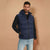 A man wearing a navy blue hooded puffer vest over a black long-sleeve shirt and light blue jeans, standing against a solid beige background.