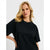 A blonde woman wearing a black oversized short-sleeve t-shirt with a gold chain necklace, photographed against a light gray studio background.