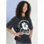 A young girl with curly dark hair wearing a black acid-wash graphic t-shirt with a white hibiscus flower and 'SUNBURG' text, posing against a light blue and white studio background.