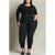 A plus-size model wearing black high-waisted skinny jeans with a matching black belt, styled with a black long-sleeve top and black studded heels, standing against a textured light gray wall.