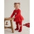 A toddler girl wearing a festive red holiday outfit with a long-sleeve top, tutu skirt, matching leggings, and knit booties, standing next to a wooden stool against a light gray background.