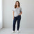 A woman modeling a light gray short-sleeve t-shirt with a small geometric cross detail at the neckline, paired with dark blue jeans and white sneakers, standing in a bright studio setting.