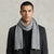 A man wearing a light gray ribbed knit scarf wrapped around his neck, styled with a dark gray blazer and black shirt against a textured light gray background.
