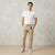 A man wearing beige drawstring jogger pants, a white t-shirt, and gray sneakers, standing against a textured light gray wall.