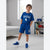 A smiling boy stands in a bright room wearing a royal blue boxing-themed t-shirt and matching shorts with white graphics, white socks, and blue sneakers.