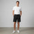 A man stands in a studio wearing a plain white short-sleeve crew-neck t-shirt, black shorts, and white sneakers against a light gray background.