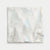 A square white fabric item, likely a napkin or small cloth square, featuring a printed pattern of shimmering silver and light blue glittery pine trees, shown flat against a light gray background.