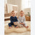 Two smiling babies sitting on a fluffy cream rug in a bright nursery. The baby on the left wears a navy blue long-sleeve one-piece and holds a beige stuffed animal, while the baby on the right wears a light gray long-sleeve one-piece.