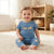 A smiling toddler sits barefoot on a cream rug in a bright room, wearing a blue short-sleeve one-piece outfit with a yellow dog graphic and paw-print details on the legs.