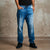 A person wearing medium-wash blue denim jeans with a faded wash, paired with a black t-shirt and black sneakers, standing against a textured concrete wall.