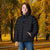 A young boy wearing a black puffer jacket and blue jeans, standing in a park with autumn trees in the background.