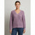 A woman wearing a textured, lavender-purple V-neck knit sweater and dark blue jeans, standing against a neutral light gray background.