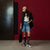 A model stands against a dark red wall wearing a beige baseball cap, a black blazer, a white button-up shirt, light-wash denim shorts, black socks, and white sneakers. She is holding a black mesh bag.