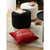 A festive Christmas throw pillow in red with gold 'Merry Christmas' lettering, styled on a cream knit rug next to two round ottomans in black and white with holiday decorations.