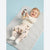 A baby lying on a light gray changing mat, wearing a cream long-sleeve top and pale pink pants with a penguin print. The baby is holding a small penguin toy against a light blue background.