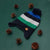 A striped knit beanie with a pom-pom, laid flat on a dark green background and surrounded by scattered pinecones. The hat has horizontal stripes in navy, blue, white, and green, with a small tan leather patch near the ear flap.