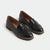A pair of glossy black patent leather penny loafers with tassel straps, displayed on a clean off-white background.