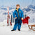 A child wearing a bright blue ski suit with yellow zipper accents stands in a snowy outdoor-style set with mountains in the background, skis, a striped chair, a red fringed blanket, and a sled.