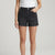 A model wearing black denim shorts with a raw, frayed hem and a white ribbed tank top, shown from the waist down against a light gray studio background.