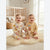 Two babies sitting on a fluffy cream rug in a bright nursery, wearing matching long-sleeve footless pajamas. The baby on the left wears a cream pajama set with colorful animal prints, and the baby on the right wears a tan/yellow pajama set with animal prints.