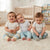 Three smiling babies sitting together on a white shag rug in a bright nursery. They are wearing matching light gray t-shirts and different pastel-colored pants: one in cream and blue striped pants, one in light blue pants, and one in gray and white patterned pants.