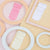 Three soft fabric baby socks in pink, white, and cream are arranged on pastel geometric paper circles and frames.