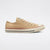 A classic low-top canvas sneaker in a light tan/beige color, featuring a cream rubber toe cap, a thin red stripe above the sole, and a navy blue stripe below it, set against a plain white background.