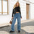 A woman stands on a cobblestone street in front of a white building, wearing a black leather moto jacket over a black crop top, high-waisted wide-leg blue jeans, and black slip-on shoes, while holding a leopard-print handbag.
