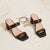A pair of black leather block-heel mule sandals with square toes and a single wide strap, displayed on a textured off-white surface next to delicate gold and black bracelets.